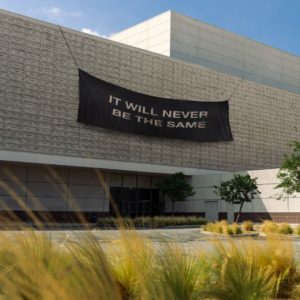 Album cover art: photo of a brick building with a black fabric banner draped over the front with the words "It Will Never Be The Same".